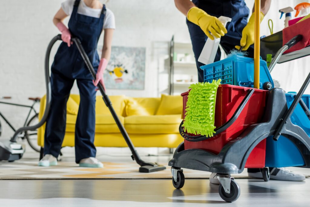 cropped view of cleaner in rubber gloves holding spray bottle near cleaning trolley and coworker