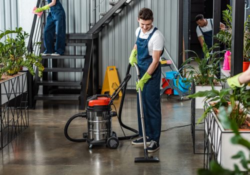 young cleaner commercial cleaning floor with vacuum cleaner near multicultural colleagues